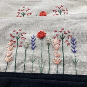 May include: Cream-colored fabric pouch with embroidered floral design. The embroidery features red, pink, purple, and white flowers with green stems. A black fabric is visible at the bottom of the image.