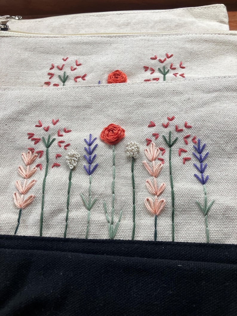 May include: Cream-colored fabric pouch with embroidered floral design. The embroidery features red, pink, purple, and white flowers with green stems. A black fabric is visible at the bottom of the image.