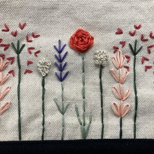 May include: Embroidered floral design featuring a central red rose and various stylized flowers in shades of pink, purple, and white. The stems are green, set against a beige fabric background. A black border is at the bottom.