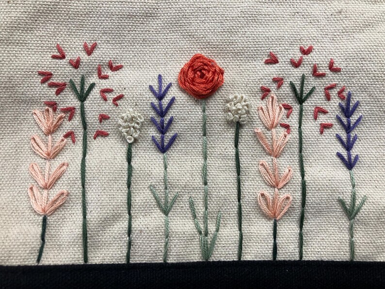 May include: Embroidered floral design featuring a central red rose and various stylized flowers in shades of pink, purple, and white. The stems are green, set against a beige fabric background. A black border is at the bottom.