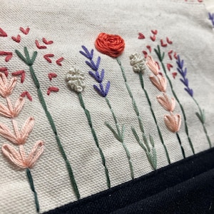 May include: Close-up of a cream-colored fabric with embroidered flowers in shades of red, pink, purple, and white. The stems are green, and the bottom edge is a solid black fabric. The embroidery is detailed and textured.