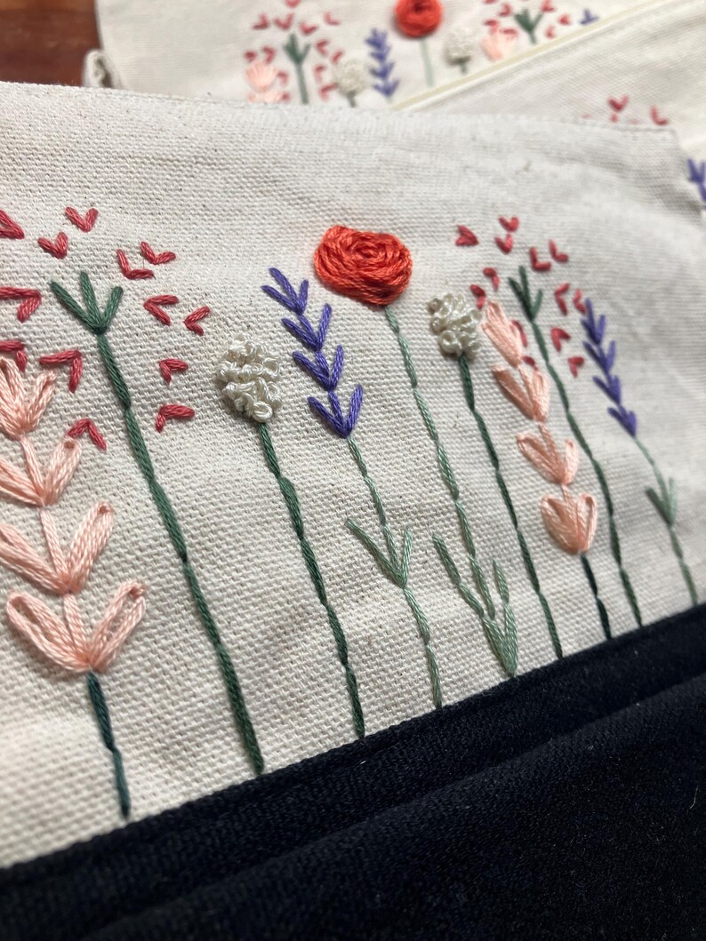 May include: Close-up of a cream-colored fabric with embroidered flowers in shades of red, pink, purple, and white. The stems are green, and the bottom edge is a solid black fabric. The embroidery is detailed and textured.
