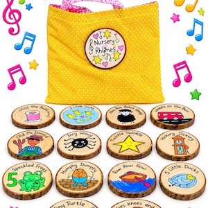 Op de afbeelding: Een gele stoffen zak met stippen en roze patroonhengsels, gelabeld Nursery Rhyme Sack 14 Songs. Het wordt vergezeld door veertien ronde houten schijven, elk voorzien van handgeschilderde illustraties en titels van klassieke kinderliedjes.