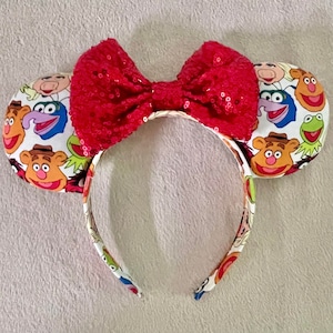 Puede incluir: Una diadema con dos orejas redondas decoradas con personajes de los Muppets. Un gran lazo rojo con lentejuelas está sujeto en la parte superior. Las orejas y la diadema son blancas con gráficos coloridos de los Muppets.