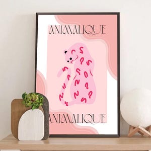 May include: A framed art print featuring a pink leopard illustration with red markings and the word "ANIMALIQUE" in a decorative font. The artwork has a soft, pastel color palette and is displayed on a wooden surface.