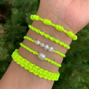 May include: A collection of neon yellow bracelets. The bracelets feature various designs, including knotted, twisted, and woven styles. Some bracelets incorporate white pearls. The bracelets are displayed on a wrist against a blurred green background.