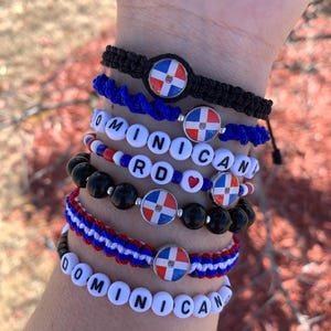 Può includere: Un set di braccialetti con la bandiera della Repubblica Dominicana. I braccialetti sono fatti di cordoncino nero, cordoncino blu, perline bianche e perline nere. I braccialetti compongono le parole "DOMINICAN" e "RD".