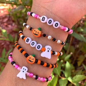May include: A collection of Halloween-themed beaded bracelets. The bracelets feature orange, black, pink, and white beads, with charms including pumpkins, ghosts, and letter beads spelling "BOO". The bracelets are designed for a festive look.
