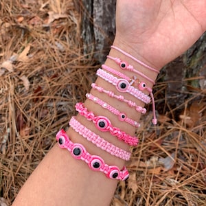 May include: A set of pink and white string bracelets with evil eye charms. The bracelets are made with different types of knots and have varying shades of pink.