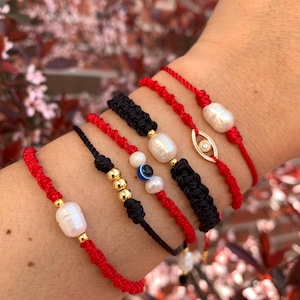 May include: A collection of handmade bracelets in red and black. Some bracelets feature pearls, gold beads, and an evil eye charm. The bracelets are knotted and braided, showcasing a variety of designs and textures.