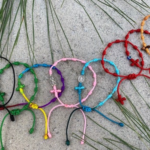 Puede incluir: Un conjunto de ocho pulseras de nudo coloridas con dijes religiosos. Las pulseras están hechas de hilo delgado y presentan una variedad de colores, incluyendo rojo, azul, verde, amarillo y rosa. Cada pulsera tiene un pequeño dije de cruz y una cuenta con una imagen religiosa.