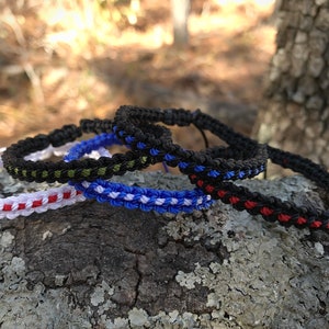 May include: Four braided bracelets in different color combinations. The bracelets are made with black, red, white, blue, and green thread. The bracelets are stacked on top of each other on a tree trunk.