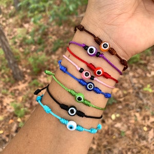 May include: Seven colorful string bracelets with an evil eye charm on each. The bracelets are in the colors brown, purple, red, pink, green, black, and blue.