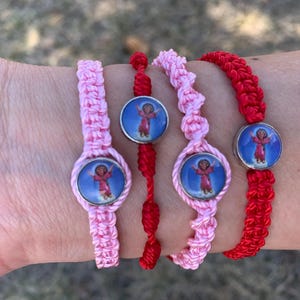 May include: Four braided bracelets with religious medallions. The bracelets are red and pink. The medallions feature a picture of a young boy with wings.