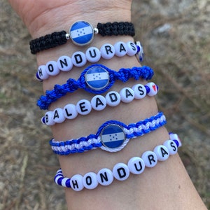 May include: Four bracelets with blue and white string and white beads spelling "HONDURAS" and "BALEADAS". Each bracelet has a charm with the Honduran flag.