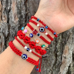 May include: A stack of red string bracelets with various blue evil eye charms, some with white accents, and a few with gold accents. The bracelets are tied with a knot and have a tassel.