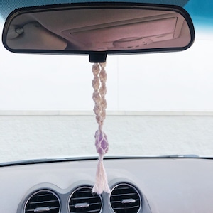 May include: A white macrame car charm with a purple amethyst crystal hanging from the rearview mirror of a car.