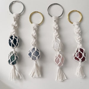 May include: Four macrame keychains with different colored stones. Each keychain has a white macrame tassel and a silver, gold, or black key ring.