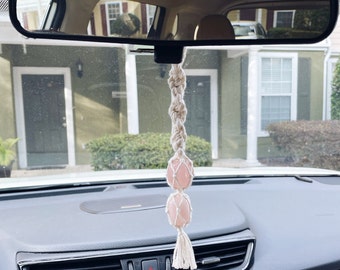 May include: A rearview mirror car charm with a macrame design and pink crystal beads. The charm is hanging from the rearview mirror of a car.