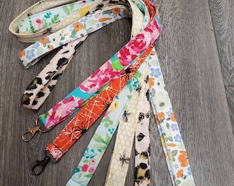 May include: A set of six colorful fabric lanyards with different patterns, including floral, bee, and spiderweb designs. Each lanyard has a gold or black metal clasp.