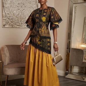 May include: A two-piece outfit consisting of a dark patterned top with flared sleeves and black lace trim, paired with a solid mustard yellow floor-length skirt. The ensemble includes a matching headwrap and a small metallic gold handbag.