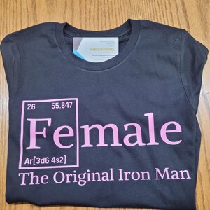 Puede incluir: Camiseta negra con la palabra "Female" en rosa, estilizada como un elemento de la tabla periódica, con el texto "The Original Iron Man" debajo. La camiseta también tiene una etiqueta que dice "Image Apparel".