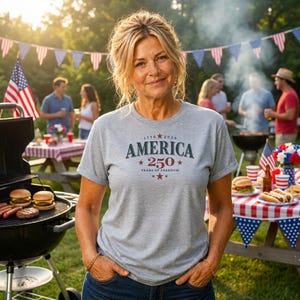 Puede incluir: Una mujer con una camiseta gris con el texto "AMERICA 250 YEARS OF FREEDOM" en azul marino y rojo. La imagen muestra una barbacoa en el jardín con hamburguesas, perritos calientes y banderas americanas.