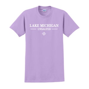 Puede incluir: Una camiseta de color lavanda con texto blanco que dice "LAKE MICHIGAN UNSALTED" y un símbolo de brújula.