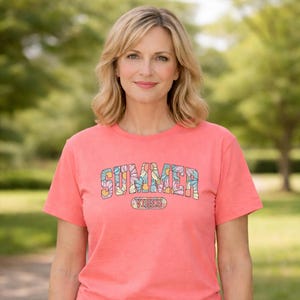 Puede incluir: Una camiseta de color coral con la palabra "SUMMER" en un diseño floral en el pecho. Debajo, la palabra "VIBES" en un diseño ovalado más pequeño. La camiseta tiene mangas cortas y cuello redondo.