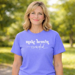Puede incluir: Una camiseta de color lila con la frase blanca "make heaven crowded". La camiseta de manga corta parece estar hecha de un material suave. La persona que lleva la camiseta sonríe frente a un fondo verde borroso.