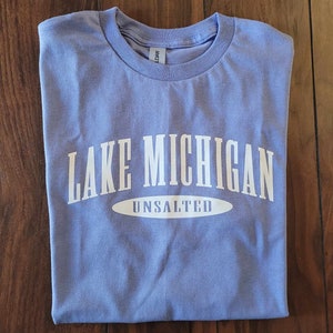 Puede incluir: Una camiseta azul claro con texto blanco que dice "Lake Michigan Unsalted".