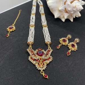 Puede incluir: Un collar de oro con un colgante rojo y blanco, un par de pendientes a juego y un pequeño adorno para el cabello rojo y dorado. El collar está hecho de cuentas blancas y tiene un diseño delicado.