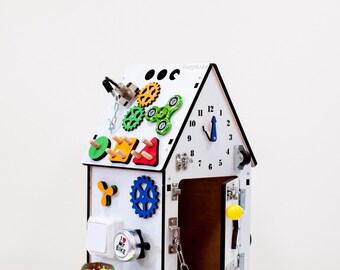 Puede incluir: Una casa de actividades de madera blanca con varios elementos interactivos coloridos. Incluye engranajes, un reloj, un spinner y una puerta con una cadena. La casa está diseñada para el juego y el aprendizaje de los niños.