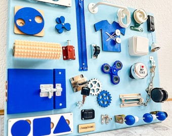 Puede incluir: Un tablero de actividades de madera azul para niños pequeños con varias texturas y actividades, que incluyen una cremallera, un pestillo de puerta, una rueda giratoria, un interruptor de luz, una campana, una llave, engranajes, un spinner, un rodillo y un clasificador de formas. El tablero está montado en una pared blanca.