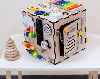 Puede incluir: Un cubo de actividades de madera con varios elementos interactivos para niños pequeños. Incluye un xilófono, engranajes, un mando a distancia, una puerta con pestillo y un laberinto de cuentas. Un juguete de anillos apilables de madera está al lado. Mide unos 25 cm.