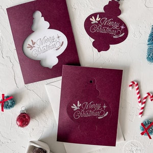 May include: Two burgundy cards with a white cutout of an ornament that says "Merry Christmas" in a script font. The ornament is decorated with small white feathers. The cards are on a white surface with other Christmas decorations.