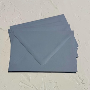 May include: A stack of five blue-grey envelopes. The envelopes are arranged in a fan shape, with the top envelope slightly overlapping the others. The paper has a matte finish. The envelopes are on a white textured surface.