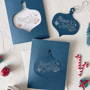 May include: Three blue cards with a white ornament cut-out. The ornament has the text "Season's Greetings" in white script. The cards are on a white surface with a red and white candy cane and a small red and white Christmas tree.