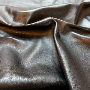 Könnte beinhalten: Nahaufnahme eines dunkelbraunen Ledermaterials mit strukturierter Oberfläche. Das Leder ist gefaltet, wodurch Schatten und Lichtreflexe entstehen, die seine geschmeidige Qualität hervorheben. Das Material scheint neu und unbenutzt zu sein.