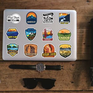 Peut inclure: Un ordinateur portable orn&eacute; d'autocollants sur le th&egrave;me du voyage, avec des motifs de parcs nationaux et des phrases comme "Find Your Wild" et "Wanderlust". Sont inclus un smartphone, une montre, des lunettes de soleil, des &eacute;couteurs et un tr&eacute;pied, dispos&eacute;s sur une surface en bois.