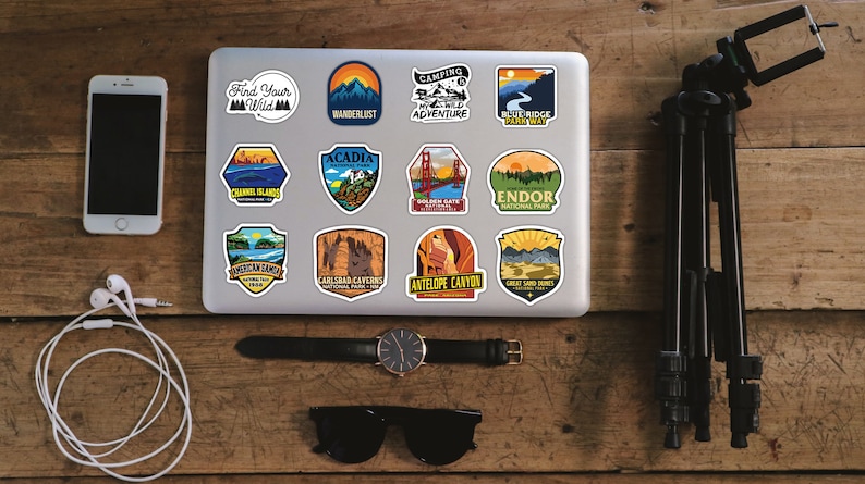Peut inclure: Un ordinateur portable orn&eacute; d'autocollants sur le th&egrave;me du voyage, avec des motifs de parcs nationaux et des phrases comme "Find Your Wild" et "Wanderlust". Sont inclus un smartphone, une montre, des lunettes de soleil, des &eacute;couteurs et un tr&eacute;pied, dispos&eacute;s sur une surface en bois.