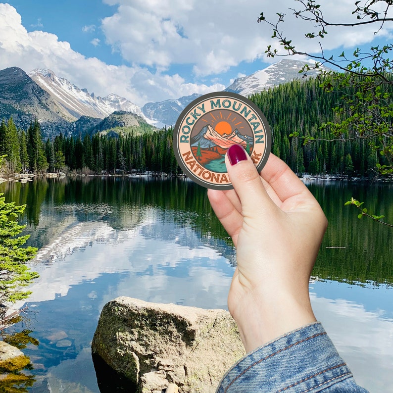 Peut inclure: Un autocollant rond repr&eacute;sentant un paysage de montagne avec le texte "Rocky Mountain National Park". Le design comprend un soleil, des montagnes et une rivi&egrave;re, avec une palette de couleurs orange, verte et marron. L'autocollant est tenu devant un lac et des montagnes.