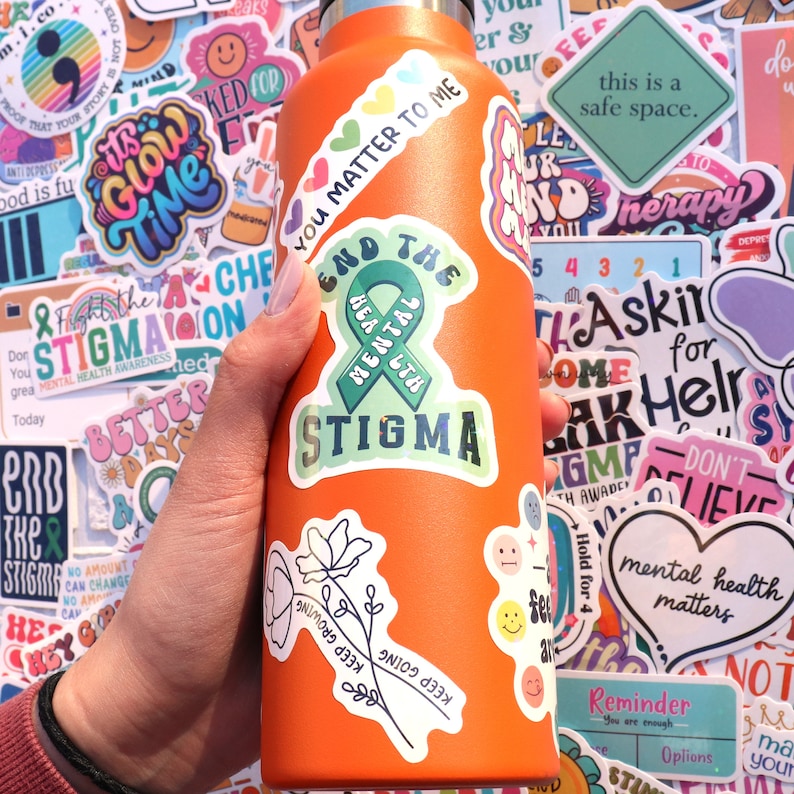 Pu&ograve; includere: Una bottiglia d'acqua in acciaio inossidabile arancione ricoperta di adesivi colorati. Gli adesivi presentano frasi come "You Matter to Me", "End the Stigma", "Keep Going" e "Mental Health Matters".