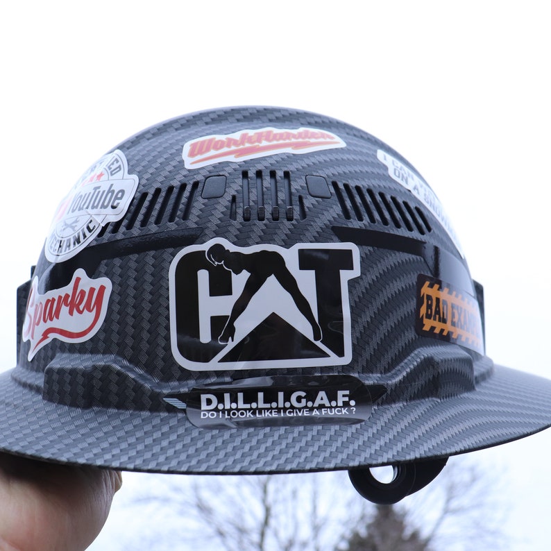 May include: A black hard hat with a carbon fiber pattern. The hard hat has several stickers on it, including a "CAT" sticker with a woman in a suggestive pose, a "D.I.L.L.I.G.A.F." sticker, and a "Work Hard" sticker.