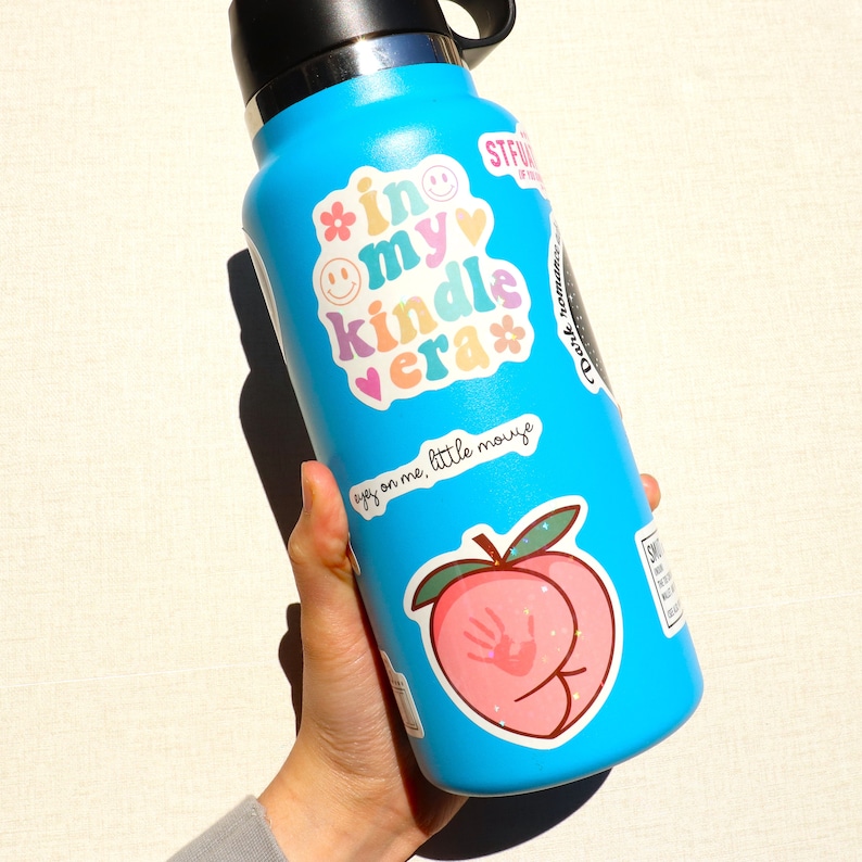 May include: A blue reusable water bottle with various stickers, including a peach with a handprint, a smiley face, a heart, and the text "in my kindle era".
