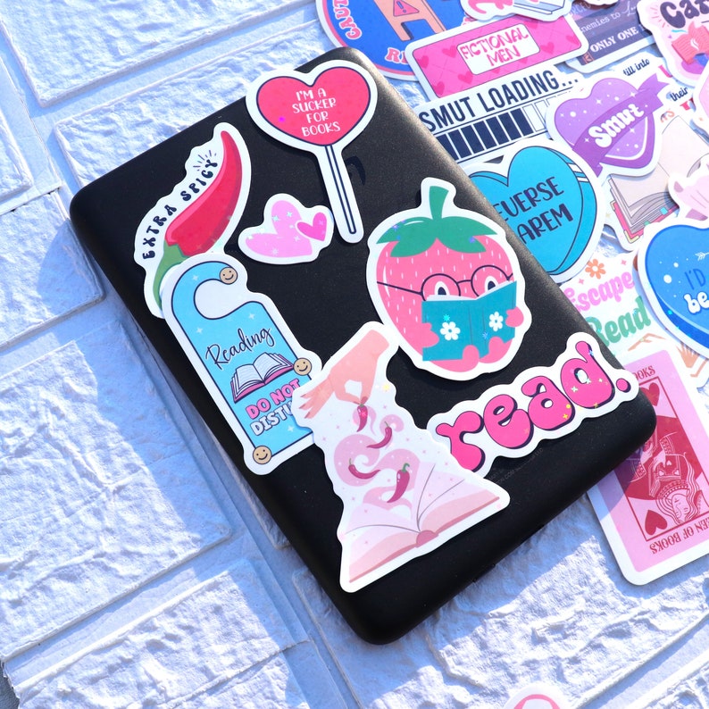 May include: A collection of colorful stickers featuring various designs, including a strawberry reading a book, a door hanger that says "Reading", a book with chili peppers, and the word "read" in pink letters. The stickers are on a black surface.
