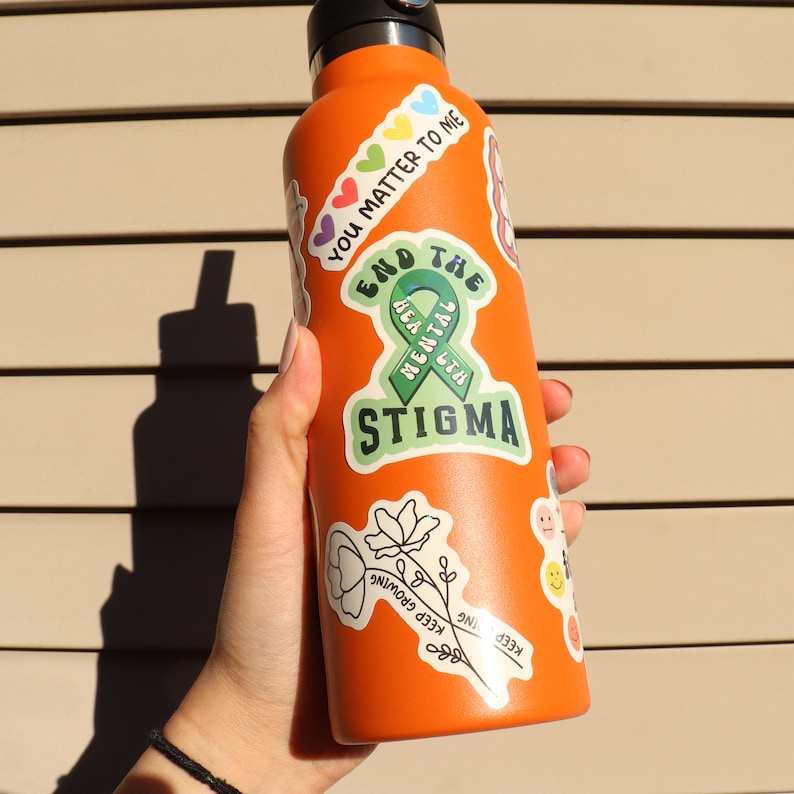 Pu&ograve; includere: Bottiglia d'acqua riutilizzabile arancione con vari adesivi, tra cui un nastro verde con il testo "End the Mental Health Stigma".
