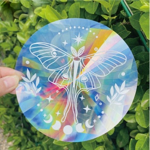 May include: A holographic sticker featuring a white outline of a moth with iridescent rainbow colors and stars. The sticker has a circular shape.