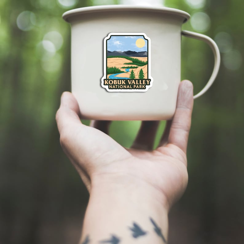 Peut inclure: Une tasse en &eacute;mail cr&egrave;me avec un autocollant repr&eacute;sentant un paysage du parc national de Kobuk Valley. L'autocollant montre une rivi&egrave;re, des montagnes, des arbres et un soleil sous un ciel bleu. La tasse a une anse et est tenue dans une main.