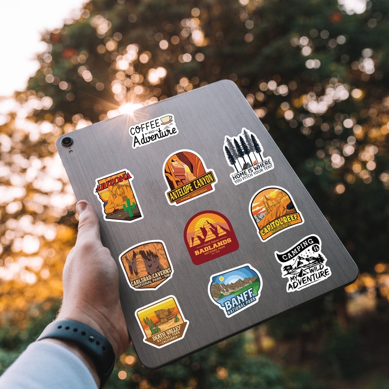 Peut inclure: Une collection d'autocollants color&eacute;s sur le th&egrave;me du voyage, coll&eacute;s sur une tablette argent&eacute;e. Les autocollants pr&eacute;sentent des dessins de parcs nationaux, de paysages et des textes tels que "Arizona" et "Camping: My Wild Adventure".
